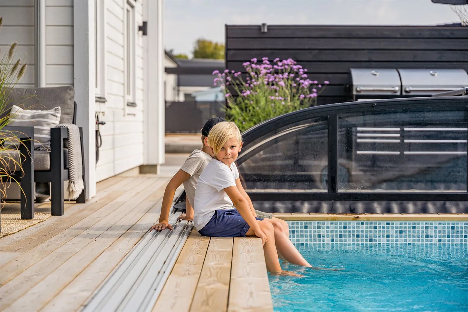Två barn på en uteplats med pool, svart insynsskydd, grill och jätteverbena i bakgrunden